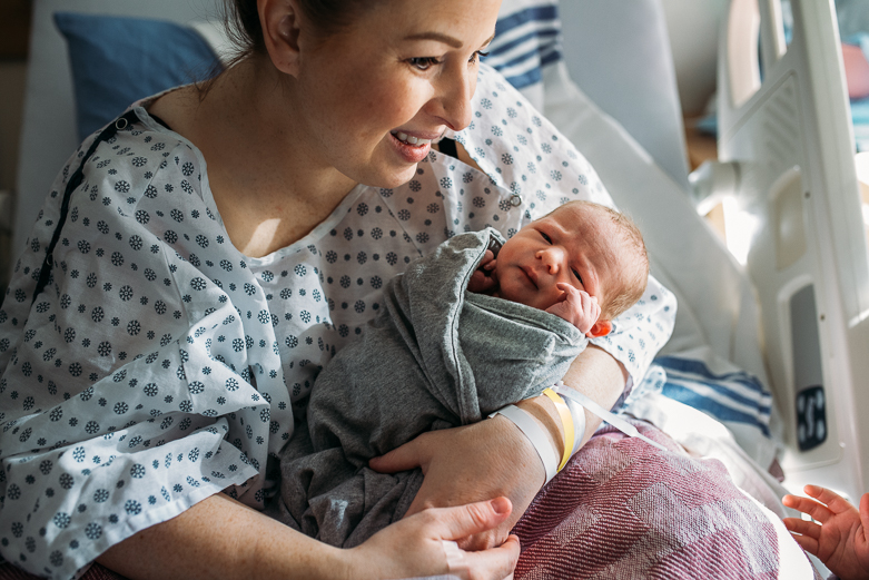 Mother in hospital gown holding newborn boy. She is looking at her young daughter not in the frame. We can see daughter's hand.